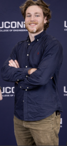 A young man in a blue button-down shirt stands in front of a wall with the UConn wordmark. 